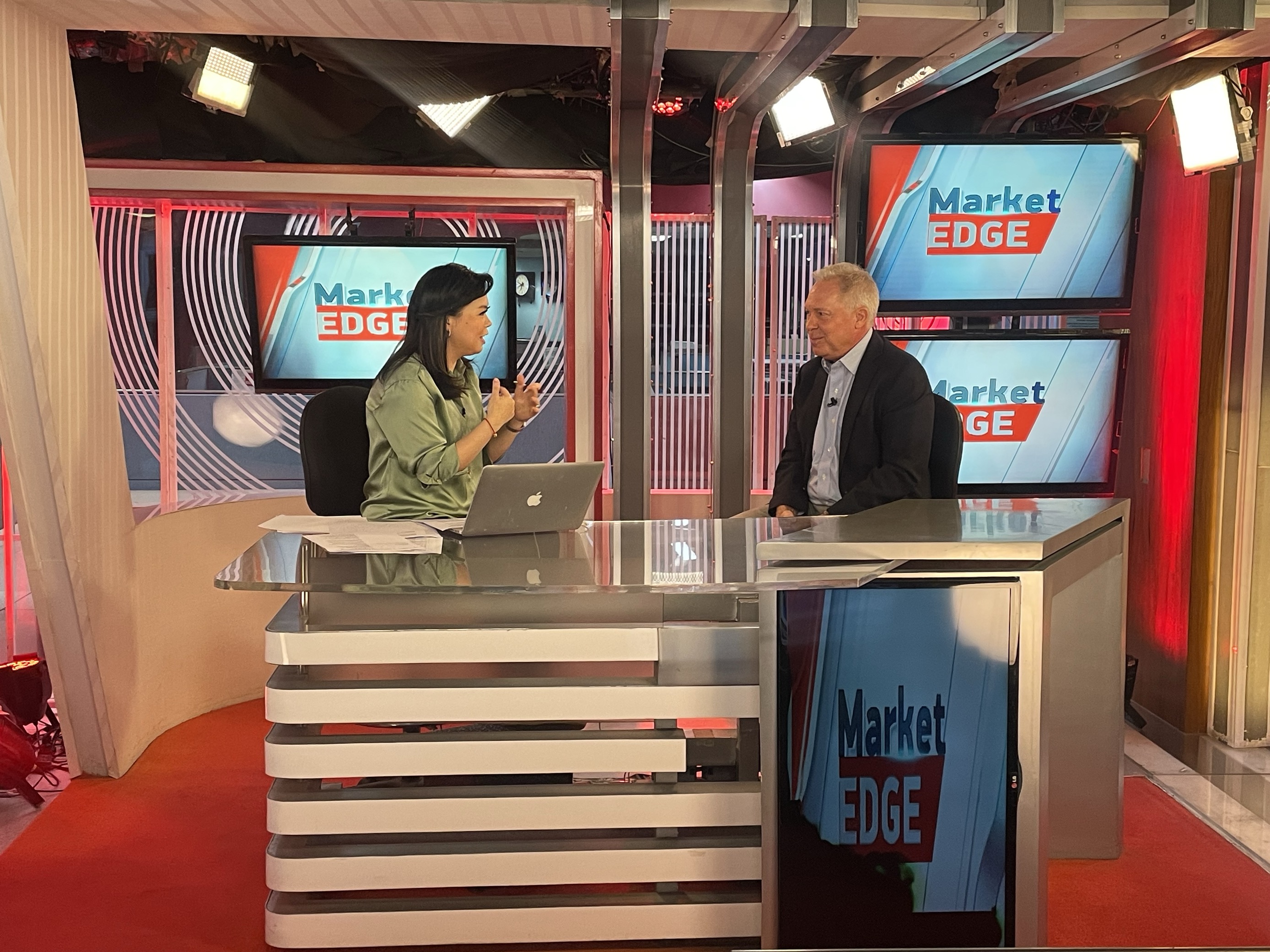 Photo from a TV studio, "Market Edge" program logo displayed on screens in the background.